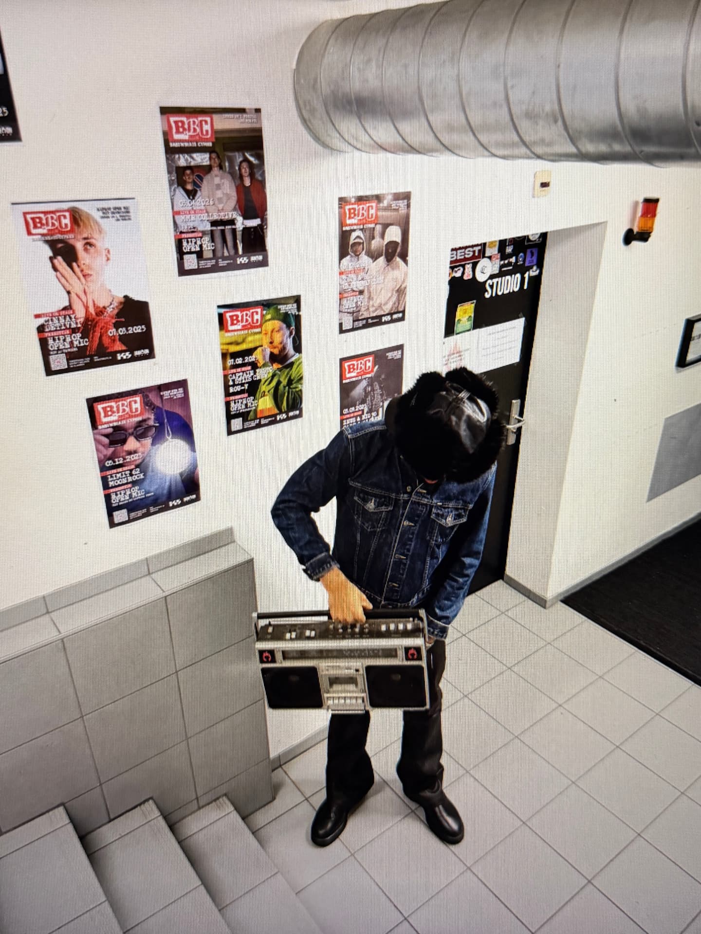 Jaywakeup holding a vintage boombox in a studio corridor lined with concert posters