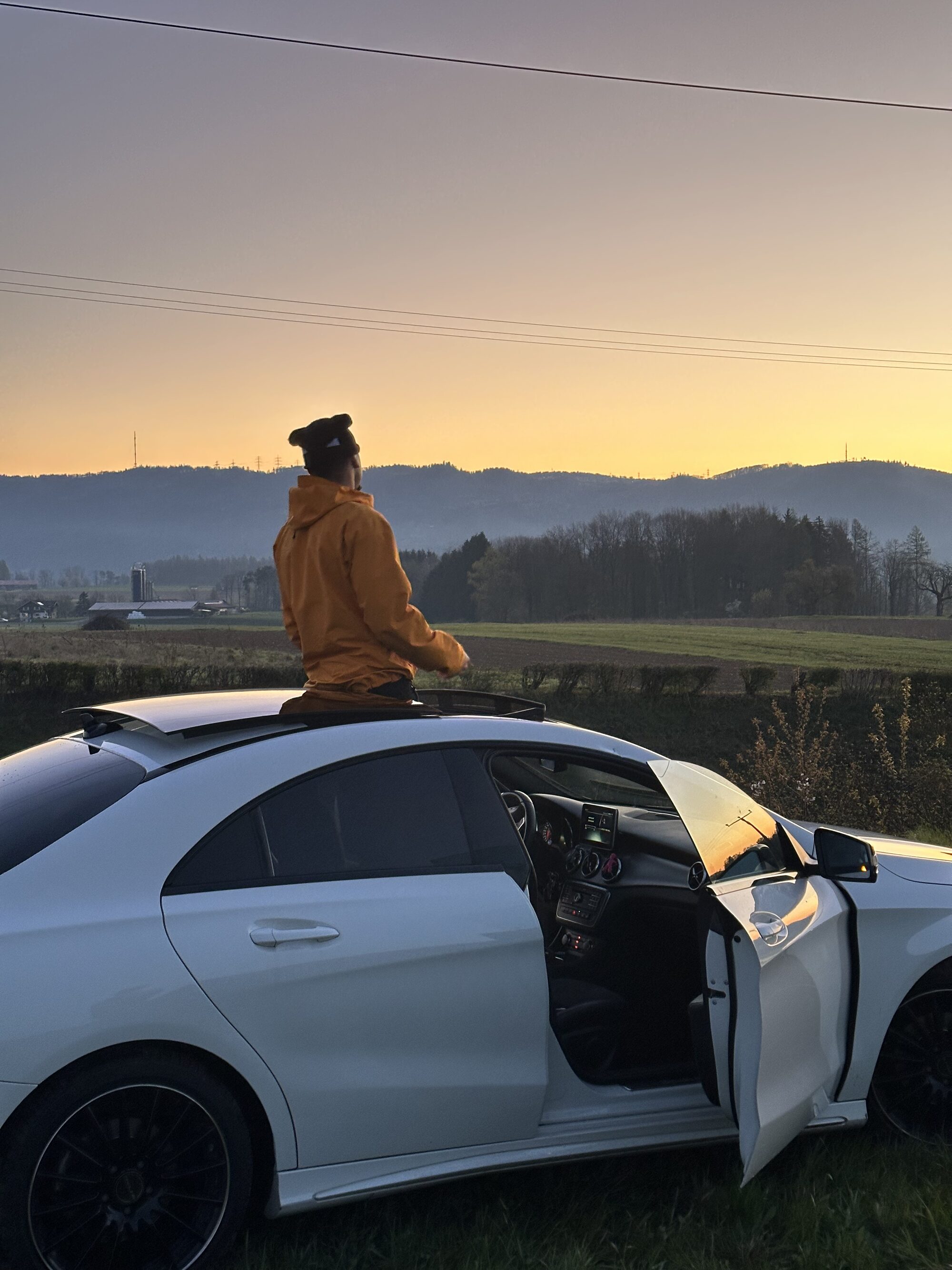 Jaywakeup in an orange hooded jacket sitting on the roof of a white car at sunset in the countryside